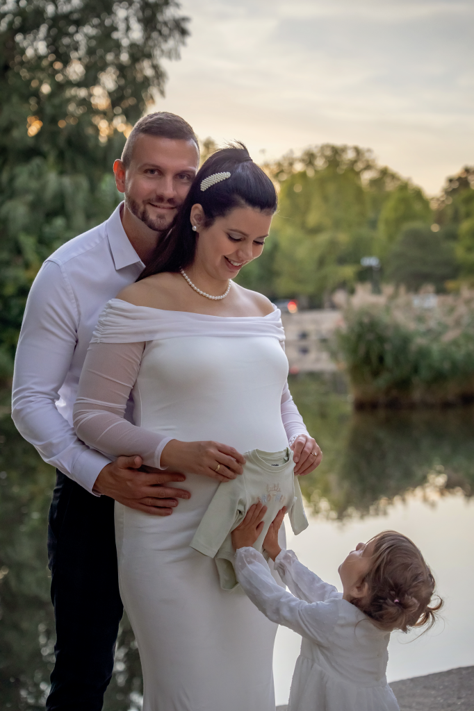 Nagytesó a kismama terhes pocakjához fogja a kisbaba bodyját, közben kispapa átöleli a kisamát.