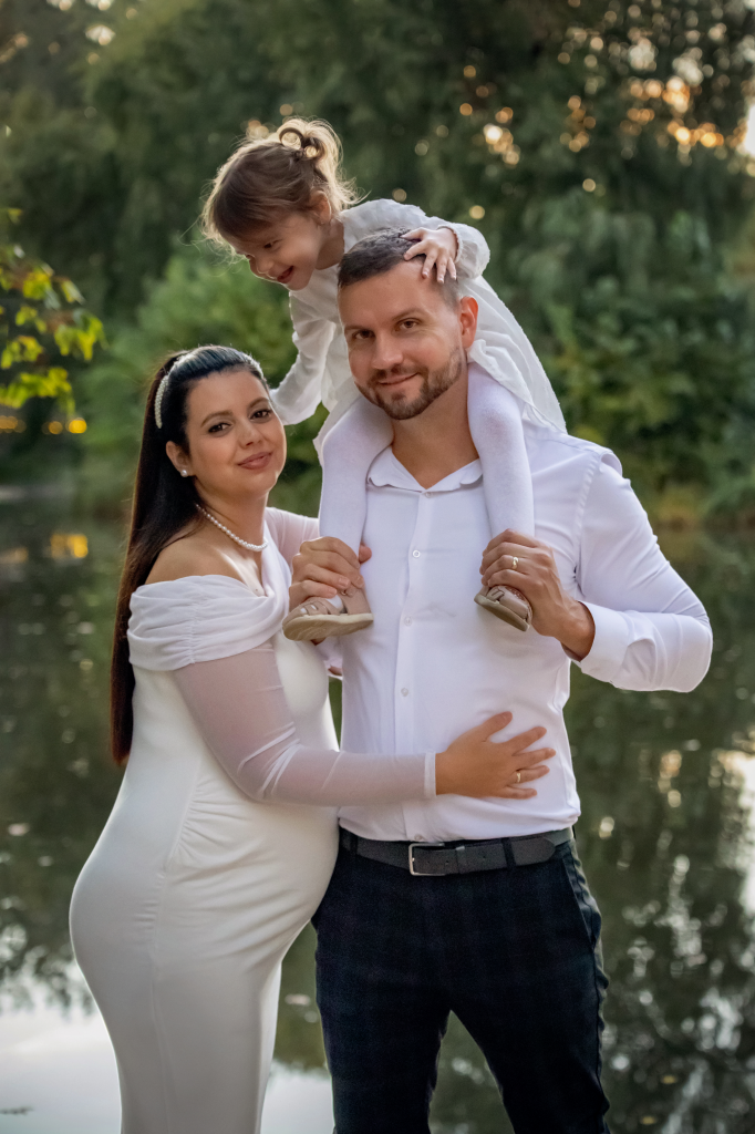 Kismama fotózás 6 Nagytesó apuka nyakában bújuk a kismamához, aki mellettük áll, és a kispapát öleli.